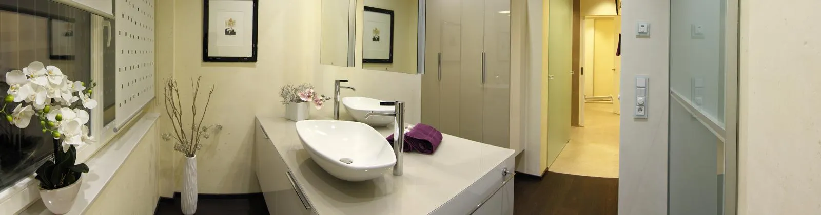 Modernes Badezimmer mit eleganter weißer Waschtischanlage, stilvollem Aufsatzwaschbecken, Spiegelwand, dekorativer Orchidee und minimalistischer Einrichtung.