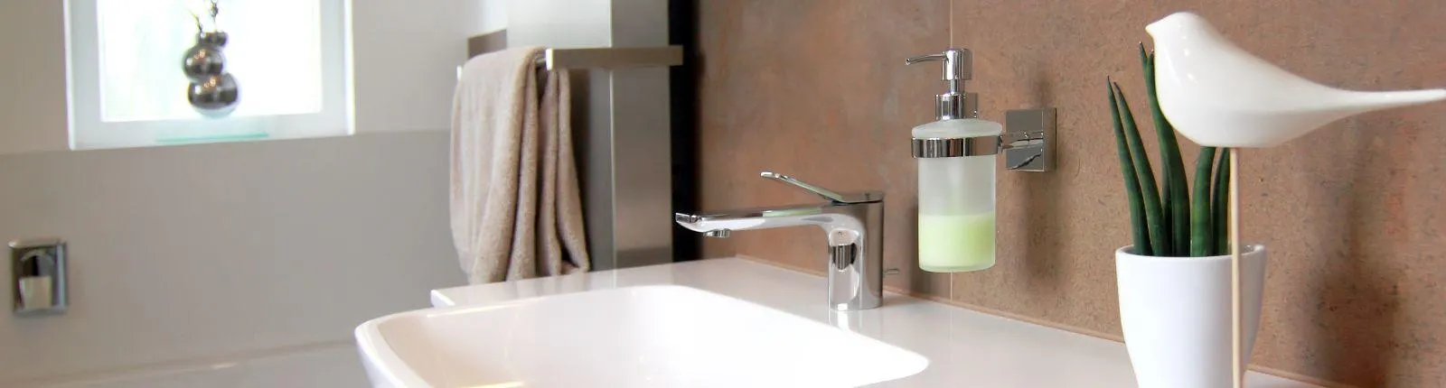 Modernes Badezimmer-Waschbecken mit verchromter Armatur, Wand-Seifenspender, dekorativer Pflanze und Keramik-Vogel im minimalistischen Design.