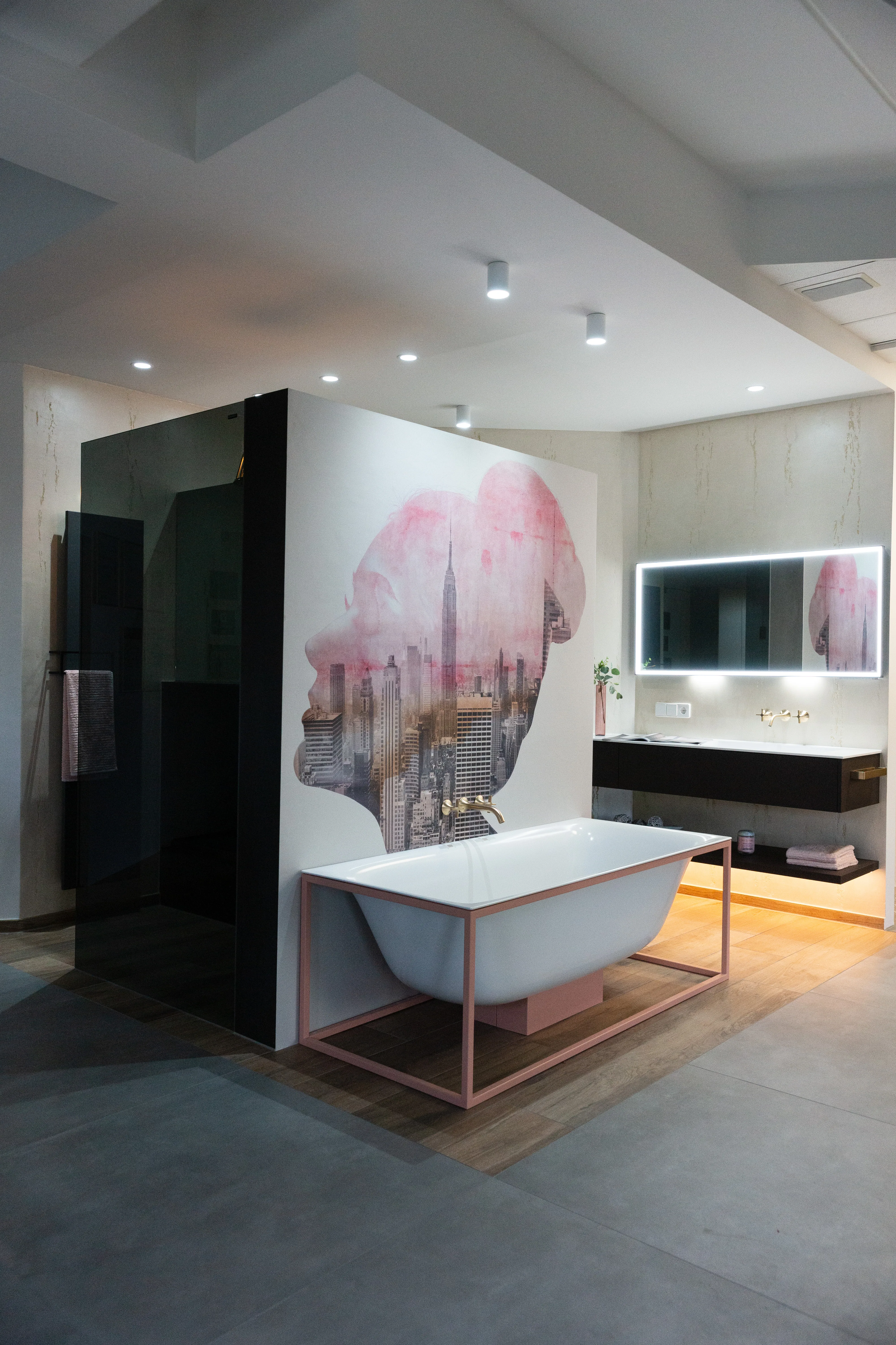 Ein modernes Badezimmer der Stübler GmbH mit freistehender Badewanne vor einer künstlerisch gestalteten Wand sowie einem beleuchteten Spiegel über einem stilvollen Waschbecken.