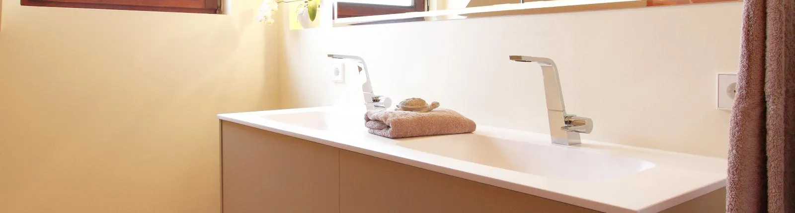 Moderner Doppelwaschtisch mit zwei verchromten Armaturen, hellem Unterschrank und gefalteten Handtüchern in einem stilvollen Badezimmer mit Naturlicht.