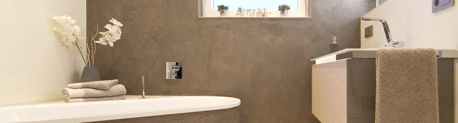 Modernes Badezimmer mit heller Badewanne, grauer Steinwand, Holzwaschtisch und minimalistischer Dekoration, darunter weiße Orchideen und Handtücher.