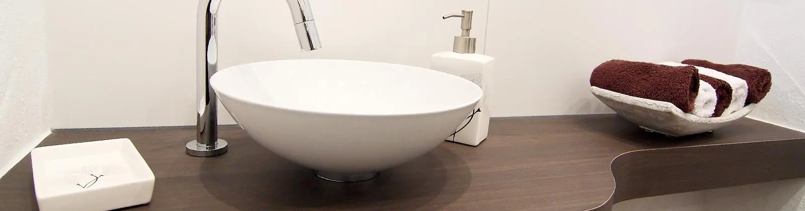 Modernes Gästebad-Waschbecken mit rundem Aufsatzbecken, schlanker Armatur, Seifenspender und dekorativen Handtüchern auf dunkler Holzablage.