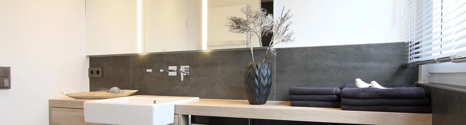 Modernes Badezimmer mit hellem Waschtisch aus Holz, eingelassenem Waschbecken, großem Spiegel und eleganter Dekoration mit schwarzen Handtüchern und Vase.