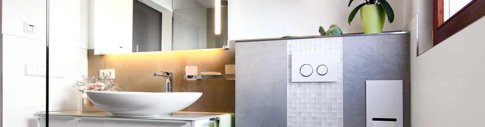 Modernes Badezimmer mit weißem Aufsatzwaschbecken, Spiegel mit Beleuchtung, grauen Fliesen und einem grünen Pflanzentopf auf dem Regal.