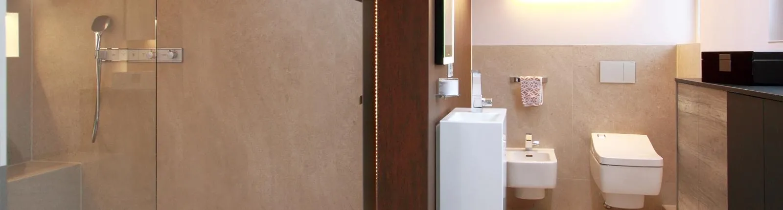 Modernes Wohnbad mit beiger Wandgestaltung, offener Dusche, wandhängendem WC und Bidet, ergänzt durch ein schmales, weißes Waschbecken und elegante Stauraumelemente.