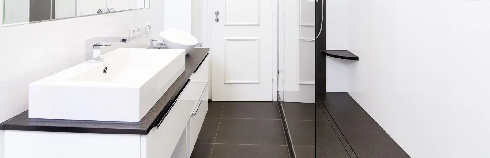 Modernes, helles Badezimmer mit weißem Waschtisch, großem Spiegel, begehbarer Dusche mit Glaswand und schwarzen Akzenten.