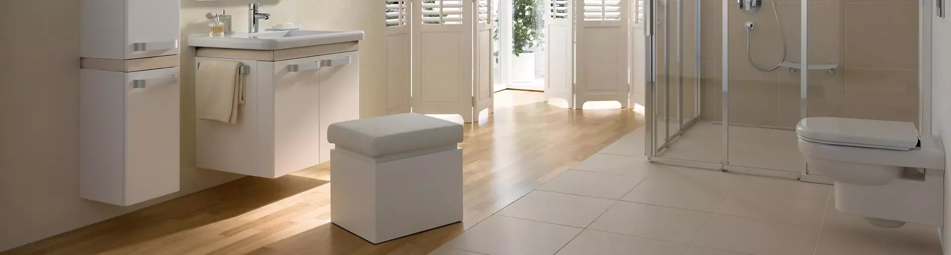 Modernes, barrierefreies Badezimmer mit bodengleicher Dusche, wandhängendem WC, Waschbecken mit Unterschrank und heller Holzfliese. Tageslicht fällt durch geöffnete Fensterläden auf den Boden.