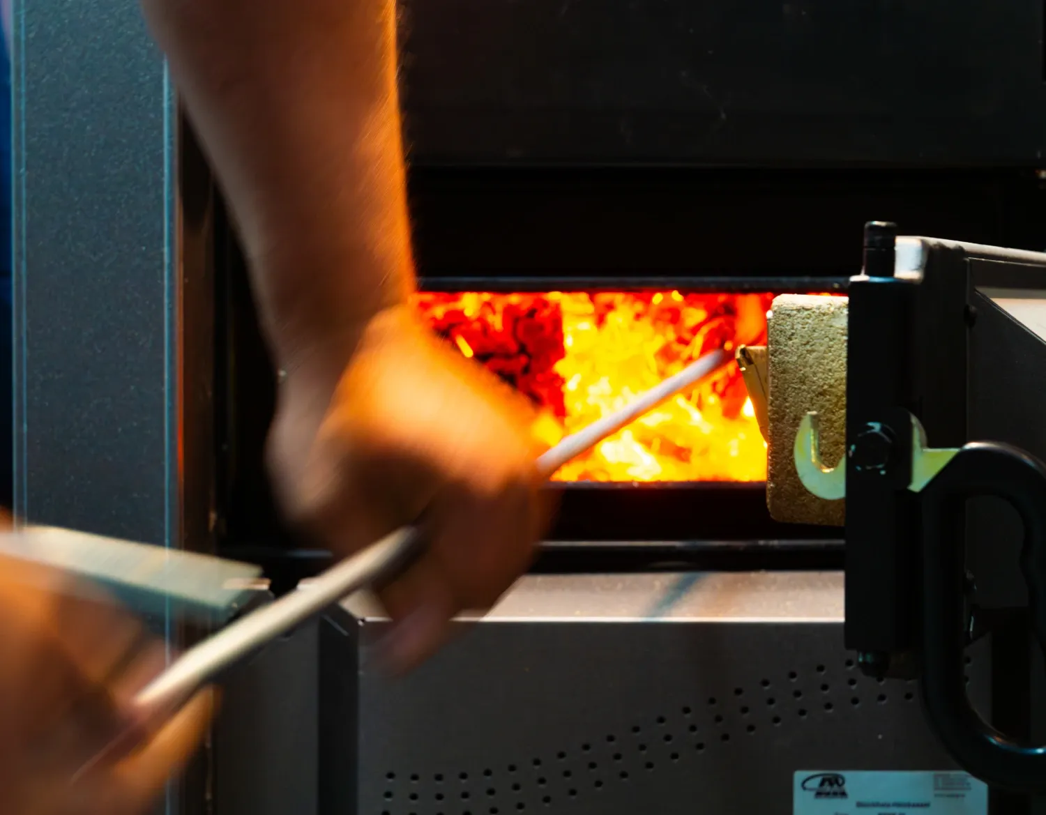 Nahaufnahme einer modernen Holzpelletheizung mit geöffneter Brennkammer und glühenden Pellets im Betrieb. Eine Person bedient die Heizungsanlage mit einem Werkzeug, während die intensive orangefarbene Glut sichtbar leuchtet.
