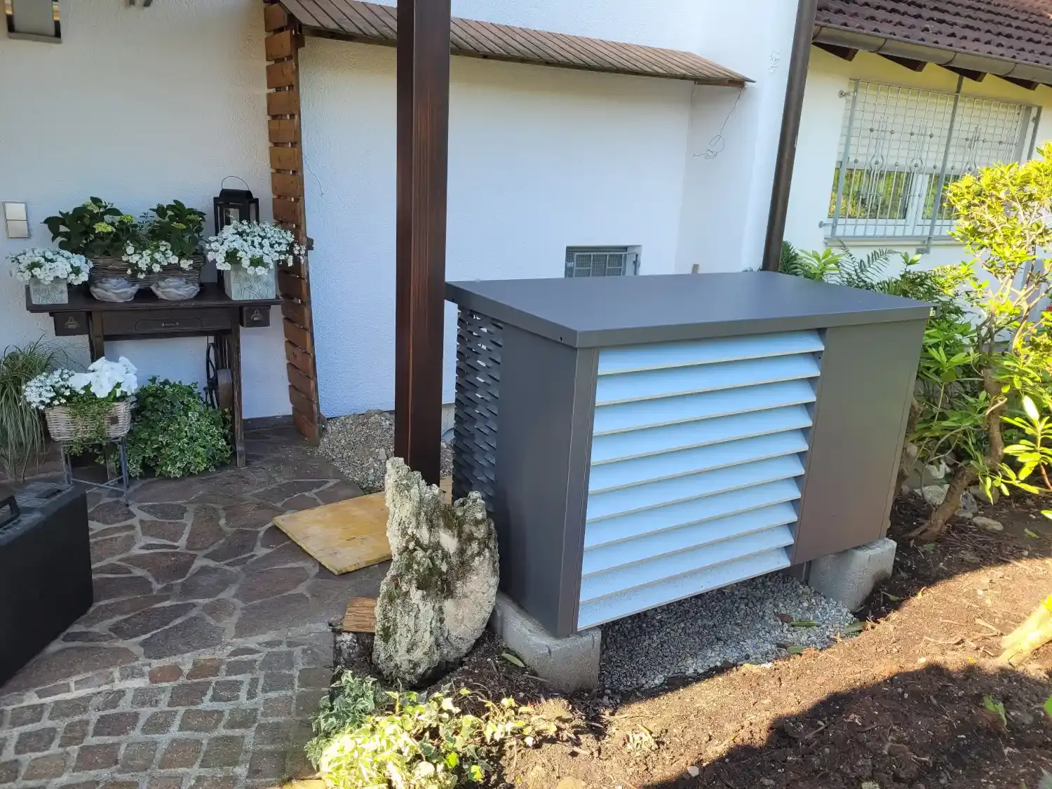 Luft-Wasser-Wärmepumpe mit Schallschutzhaube in einem gepflegten Garten neben einer Terrasse. Die schallgedämmte Außeneinheit steht auf einem Fundament und fügt sich unauffällig in die Gartenlandschaft ein.
