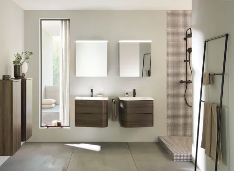 Stilvolles Bad mit hängenden Holz-Badezimmermöbeln der Serie badu von burgbad sowie Doppelwaschbecken und beleuchteten Spiegeln. Eine bodenebene Dusche mit Mosaikfliesen ergänzt das Design.