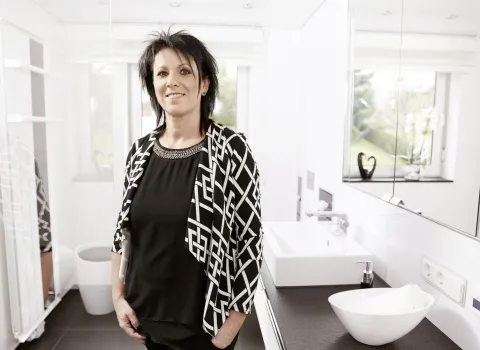 Zufriedene Kundin steht in einem modern sanierten Badezimmer mit weißem Waschtisch, Aufsatzwaschbecken, großem Spiegel und eleganter Schwarz-Weiß-Gestaltung.