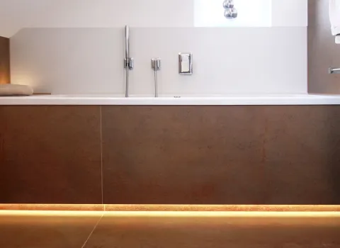 Badewanne mit moderner Wandverkleidung in Natursteinoptik, eleganten Armaturen und stimmungsvoller LED-Beleuchtung im Sockelbereich.