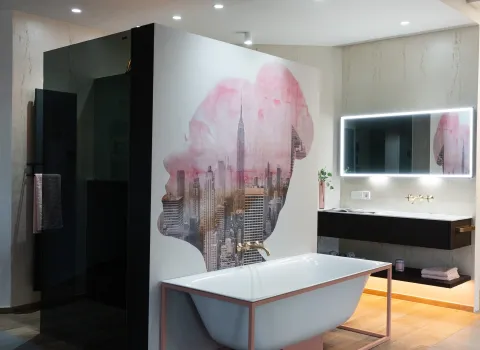 Ein modernes Badezimmer der Stübler GmbH mit freistehender Badewanne vor einer künstlerisch gestalteten Wand sowie einem beleuchteten Spiegel über einem stilvollen Waschbecken.