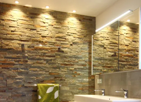 Badezimmer mit beleuchteter Natursteinwand, großem Spiegel und Doppelwaschtisch, ergänzt durch dezente Einbauleuchten und grüne Handtuch-Akzente.