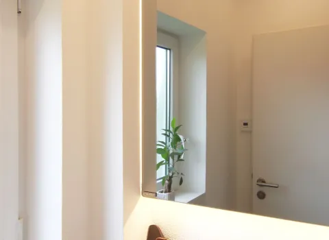 Modernes Gäste-WC mit großem beleuchtetem Spiegel, Fenster mit kleiner Pflanze auf der Fensterbank und schlichter weißer Tür im minimalistischen Design.