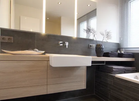 Modernes Junggesellenbad mit Holz-Waschtisch, rechteckigem Waschbecken, großem Spiegel und grauen Fliesen, ergänzt durch dekorative Accessoires.