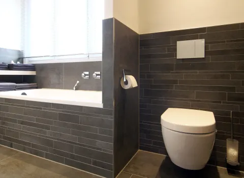 Modernes Junggesellenbad mit dunklen Fliesen, integrierter Badewanne und wandhängendem WC in klarer, minimalistischer Gestaltung.