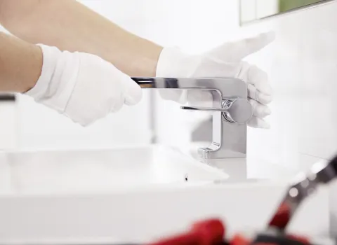 Nahaufnahme einer gloved Hand, die eine moderne verchromte Waschbeckenarmatur montiert, im Hintergrund ein weißes Waschbecken und Werkzeug im Vordergrund.