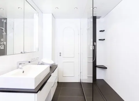 Modernes, helles Badezimmer mit großem rechteckigen Waschbecken auf dunkler Ablagefläche, Spiegelwand mit Beleuchtung und bodengleicher Glasdusche mit schwarzem Akzentstreifen, Regendusche und Eckablagen.