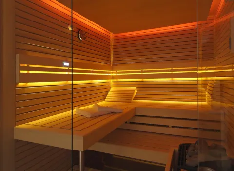 Moderne Sauna mit Holzbänken, stimmungsvoller gelb-orangefarbener Beleuchtung, Handtüchern auf der Bank und einem kleinen Saunaofen.