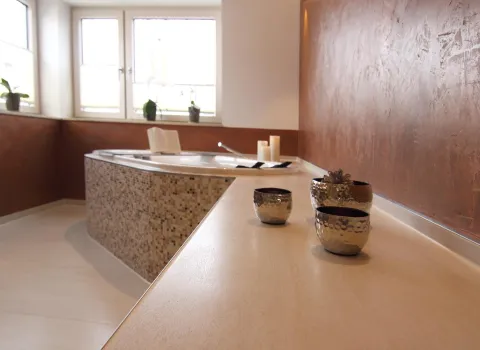 Detailansicht eines modernen Badezimmers mit dekorativen Metallschalen auf einer Ablage und einer eleganten Eckbadewanne mit Mosaikfliesen im Hintergrund.