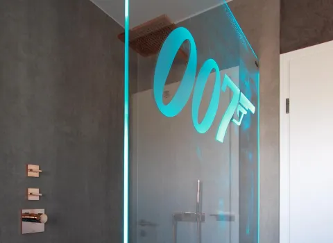 Moderne Dusche mit grauer Steinoptik-Wand, kupferfarbenen Armaturen und Glasabtrennung mit leuchtendem 007-Logo.