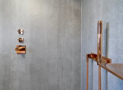 Edle Dusche mit kupferfarbenen Armaturen und Handbrause vor grauer Steinoptik-Wand, mit integrierten Bedienelementen und Ablage.
