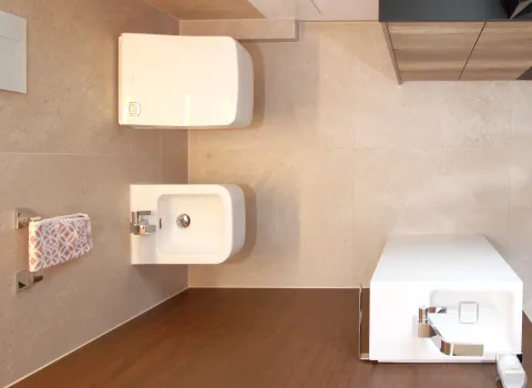 Vogelperspektive auf ein modernes Wohnbad mit wandhängendem WC, Bidet und schmalem Waschbecken, gestaltet in hellen Natursteinfliesen mit warmen Holzelementen.