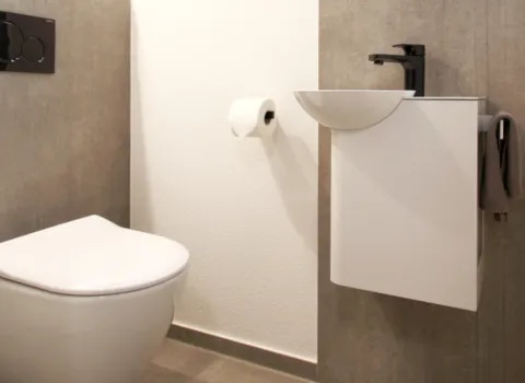 Kompaktes Gäste-WC mit wandhängender Toilette, schwarzer Betätigungsplatte, kleinem weißen Waschbecken mit schwarzer Armatur und seitlichem Handtuchhalter.