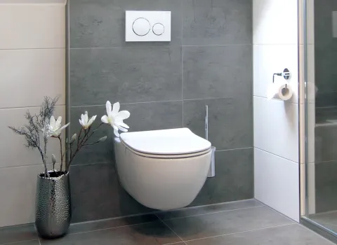 Wandhängendes WC im altersgerechten Dachbad mit grauen Fliesen und dekorativer Vase mit weißen Blumen.