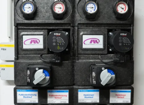 echnische Heizungssteuerung mit Pumpengruppen, Thermometern und Kupferrohren für Fußbodenheizung und Heizkörper. Die Anlage zeigt Vorlauf- und Rücklaufleitungen einer modernen Heizungsverteilung im Technikraum.