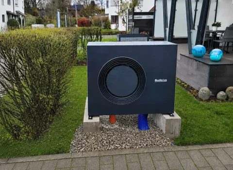 Außengerät einer modernen Heizungsanlage mit Wärmepumpe im Gartenbereich eines Neubaus. Umgeben von Grünflächen, Hecken und einer Terrasse fügt sich das System harmonisch in die Wohnumgebung ein.