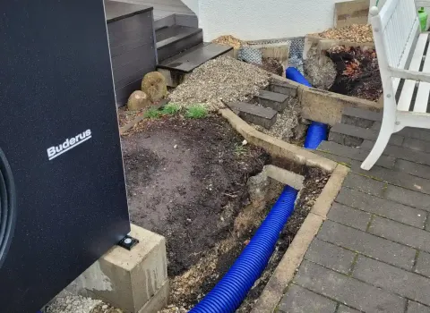 Erdarbeiten für eine Luft-Wasser-Wärmepumpe im Vorgarten eines Einfamilienhauses. Sichtbar sind geöffnete Gräben mit blauen Schutzrohren für die Leitungsführung zwischen Außeneinheit und Haustechnik.