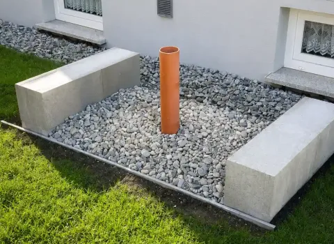 Vorbereitetes Fundament für eine Luft-Wasser-Wärmepumpe neben einem Wohnhaus. Das Kiesbett mit Leerrohr und massiven Betonfundamenten dient als Basis für die spätere Installation der Außeneinheit.