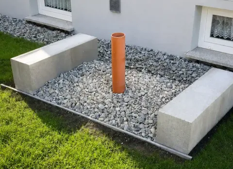 Vorbereitetes Fundament für eine Luft-Wasser-Wärmepumpe neben einem Wohnhaus. Das Kiesbett mit Leerrohr und massiven Betonfundamenten dient als Basis für die spätere Installation der Außeneinheit.
