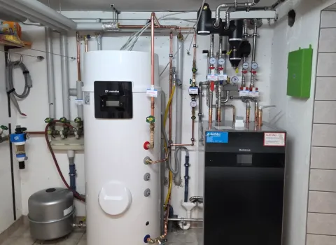Kompakte Heizungszentrale mit Wärmepumpen-Inneneinheit, Warmwasserspeicher und hydraulischen Anschlüssen. Die Anlage ist platzsparend im Keller installiert und ermöglicht energieeffizientes Heizen sowie die Warmwasserbereitung.