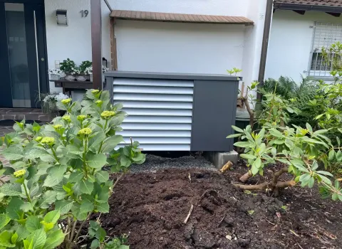 Moderne Wärmepumpen-Außeneinheit mit schallgedämmter Verkleidung im Eingangsbereich eines Wohnhauses. Die Anlage steht auf einem Fundament und fügt sich harmonisch in die Garten- und Beetgestaltung ein.
