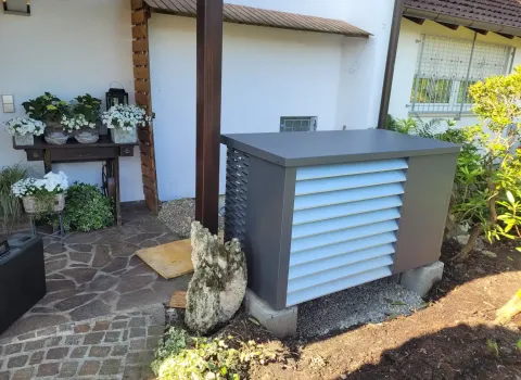 Schallgedämmte Luftwärmepumpe auf einem Kiesfundament neben Terrasse und Gartenbepflanzung. Das wetterfeste Gehäuse reduziert Betriebsgeräusche und unterstützt eine umweltfreundliche Heiztechnik für Wohngebäude.