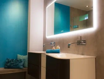 Ein modernes Badezimmer mit türkisfarbener Wand, beleuchtetem Spiegel, zwei Waschbecken mit Wandarmaturen und einer Sitzbank mit dekorativen Kissen.