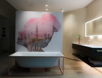 Ein stilvolles Badezimmer mit freistehender Badewanne vor einer Wand mit künstlerischem Wandbild sowie einem beleuchteten Spiegel über einem modernen Waschbecken des Unternehmens Stübler GmbH.