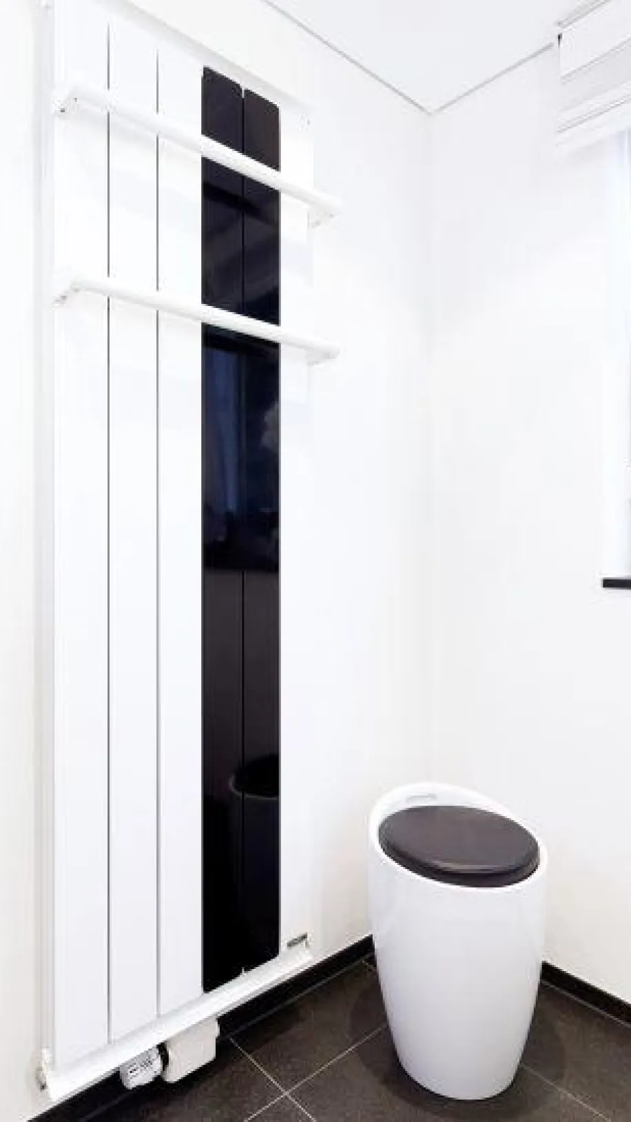 Modernes Badezimmer mit vertikalem Heizkörper in Weiß und Schwarz mit Handtuchhaltern, daneben ein weißes WC mit dunklem Sitz, vor einem Fenster mit heller Jalousie.