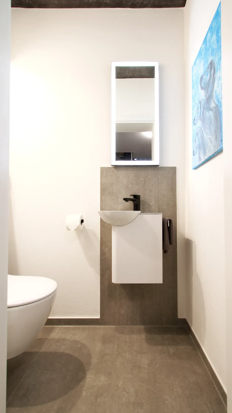 Modernes Gäste-WC mit wandhängender Toilette, kleinem Waschbecken mit schwarzer Armatur, beleuchtetem Spiegel und Wandbild.