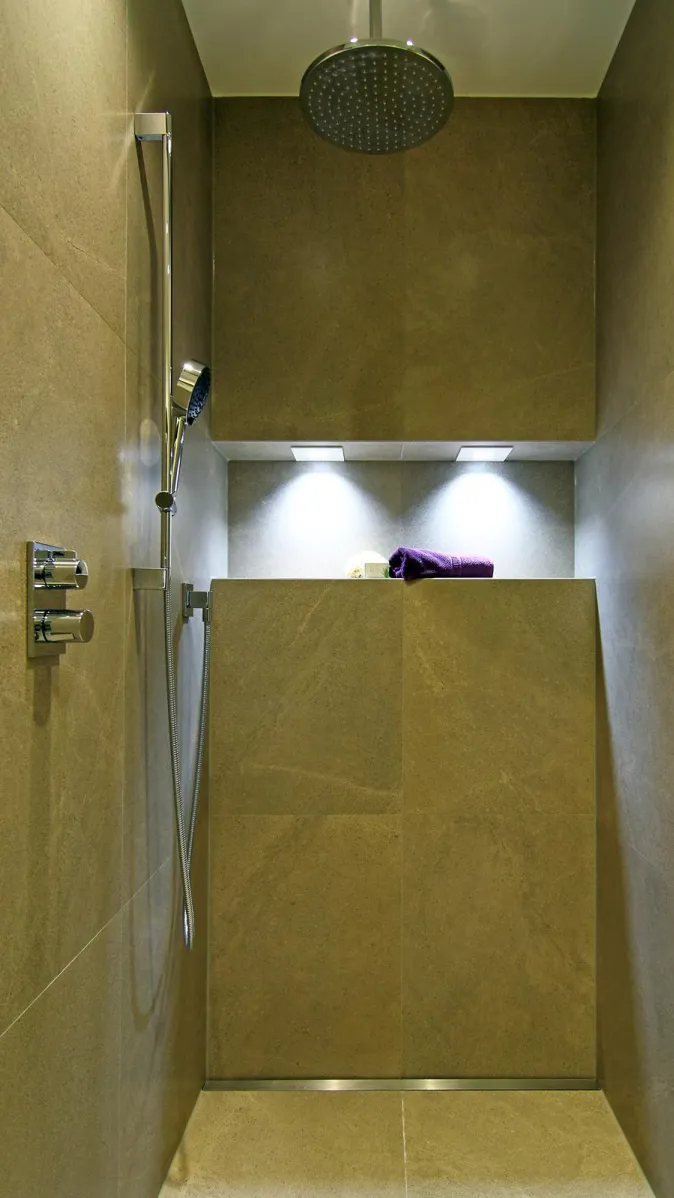 Moderne, bodengleiche Dusche mit großformatigen Natursteinfliesen, Regendusche, Handbrause, integrierter Nische mit Beleuchtung und dekorativem Handtuch.