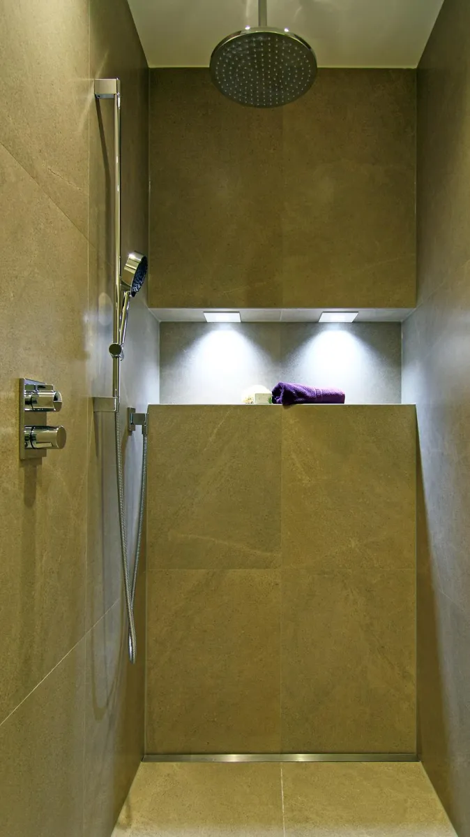 Moderne, bodengleiche Dusche mit großformatigen Natursteinfliesen, Regendusche, Handbrause, integrierter Nische mit Beleuchtung und dekorativem Handtuch.