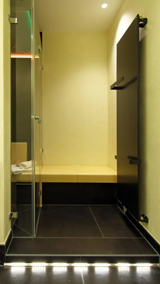 Modernes Dampfbad mit heller Sitzbank, Glasfront, dunklem Fliesenboden und dezenter LED-Beleuchtung am Bodenrand.