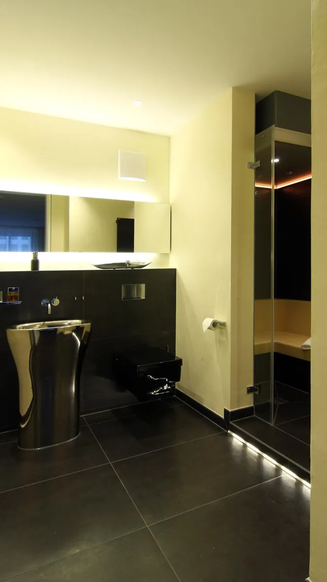 Elegantes Gästebad mit modernem Edelstahl-Waschtisch, großem Leuchtspiegel, schwarzen Bodenfliesen und angrenzendem Dampfbad mit Glasfront.