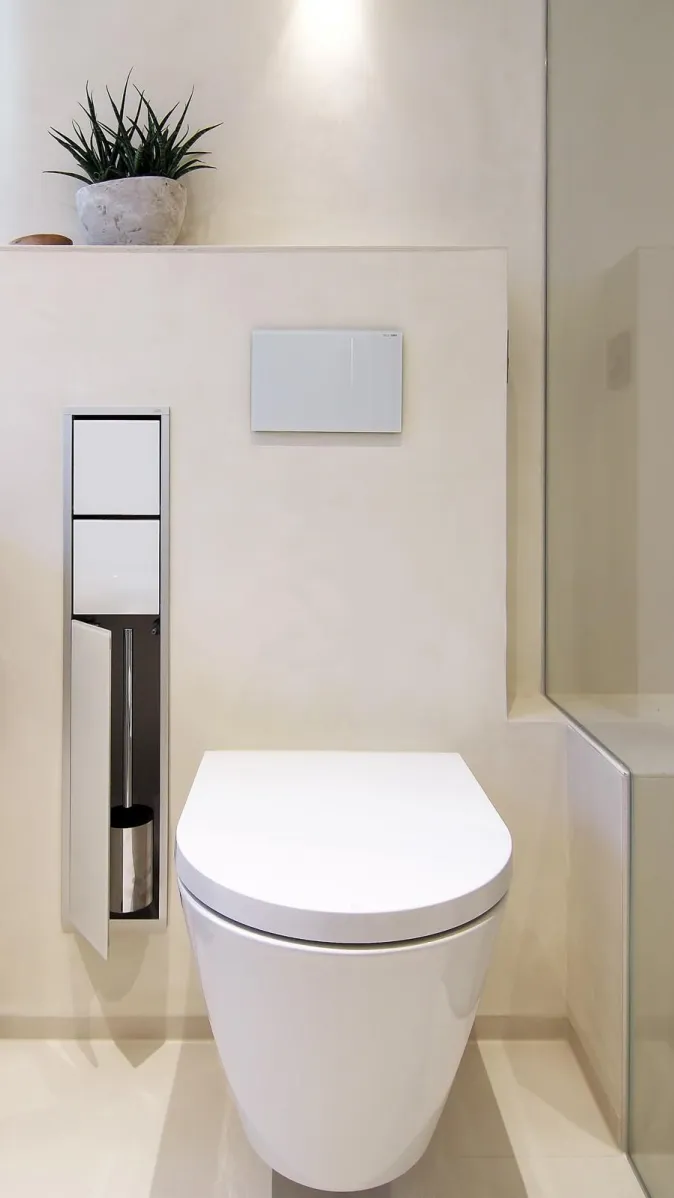 Wandhängendes WC in modernem Badezimmer mit integrierter WC-Bürste, Glasabtrennung und dekorativer Zimmerpflanze.