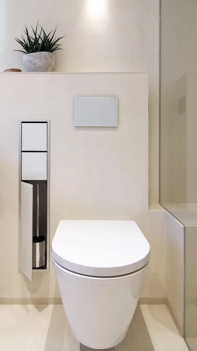 Wandhängendes WC in modernem Badezimmer mit integrierter WC-Bürste, Glasabtrennung und dekorativer Zimmerpflanze.