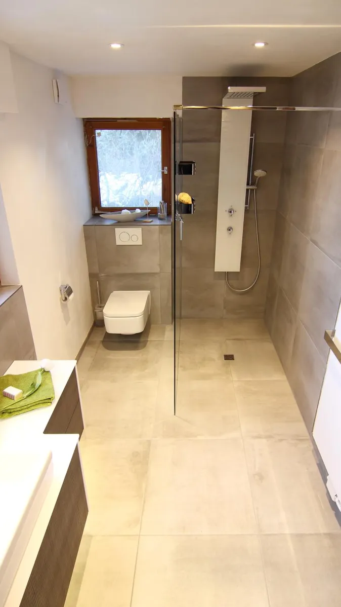 Helles Familienbad mit bodengleicher Glasdusche, wandhängendem WC und großen grauen Fliesen, ergänzt durch moderne Einrichtung und grüne Handtuch-Akzente.