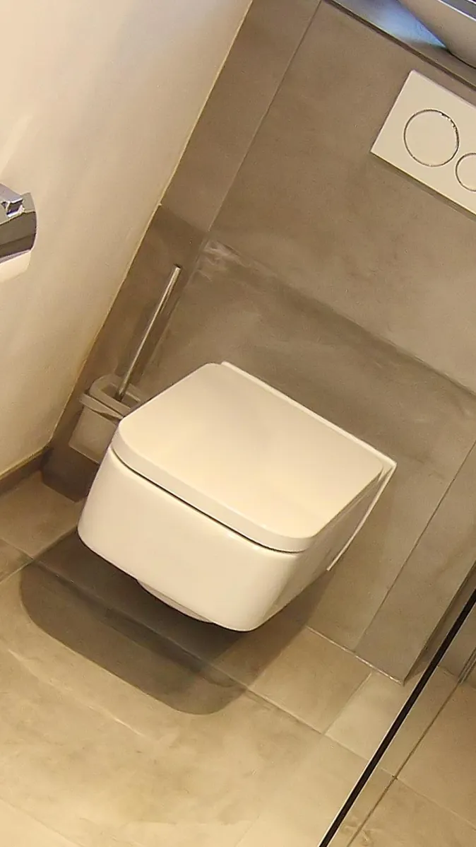 Modernes wandhängendes WC mit weißer Betätigungsplatte und großen grauen Wand- und Bodenfliesen.